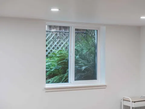 Double window with white trim in a finished basement bedroom in Seattle, WA, with ferns outside