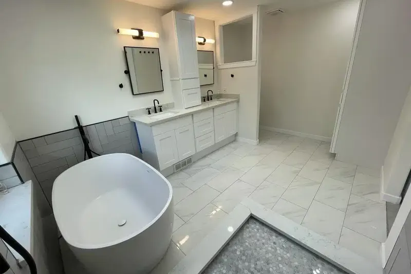 Remodeled primary bathroom in Bothell, WA with a white freestanding tub, marble look floor tile and a white shaker double vanity with black mirrors