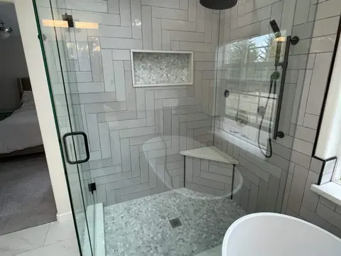 Frameless glass shower in Bothell, WA with herringbone wall tile, a corner bench, a pebble floor and a black handheld shower bar