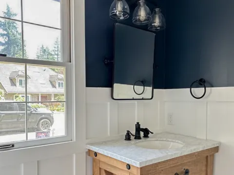Powder room in Sammamish, WA with navy walls, white wainscot, a pivot mirror and a wood vanity with a marble top beside a window
