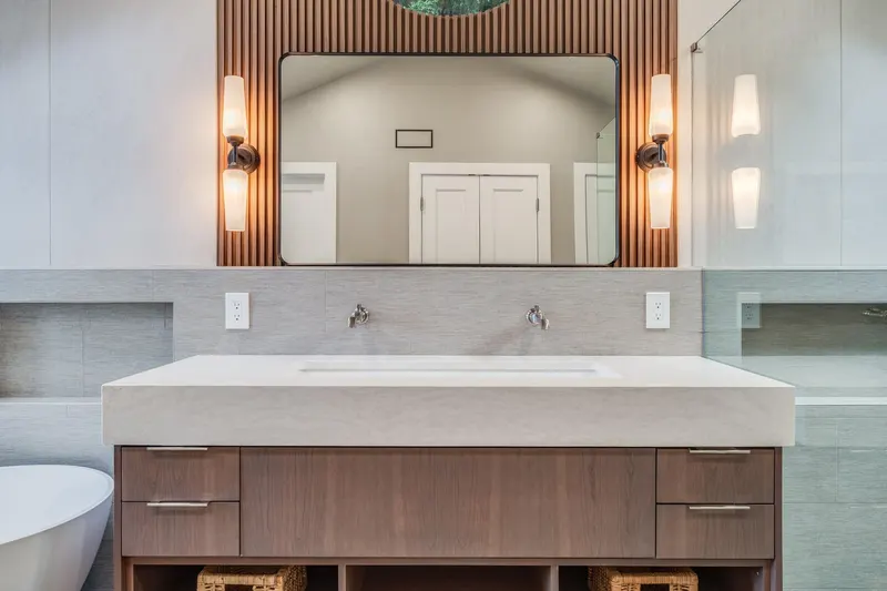 Walnut floating vanity with a wide stone sink, wall mounted faucets and slatted wood behind the mirror in a Redmond, WA primary bathroom
