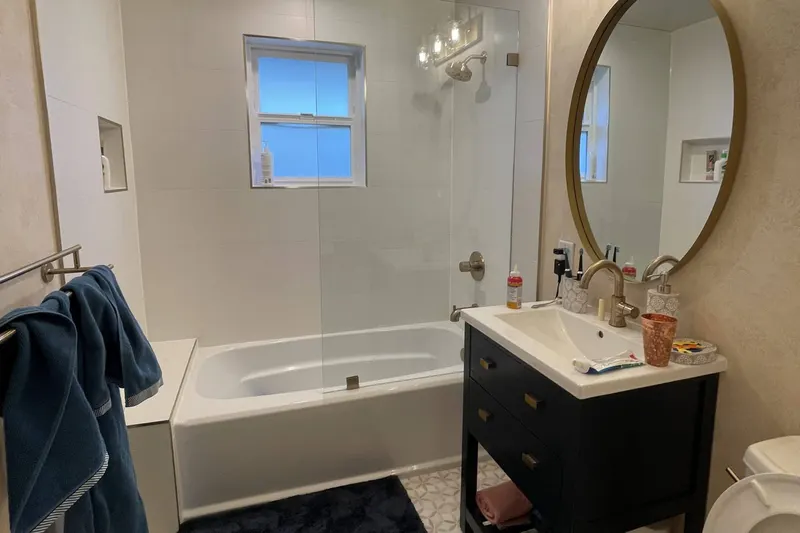Bathtub with a glass panel, a navy vanity and a round brass mirror in a guest bathroom remodel in Seattle, WA