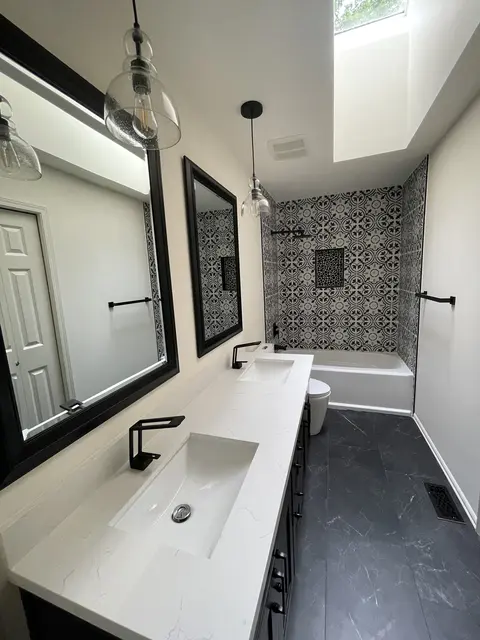 Remodeled bathroom with a long white counter on dark cabinets, glass pendant lights, black mirrors and a skylight, with the patterned tile tub at the end