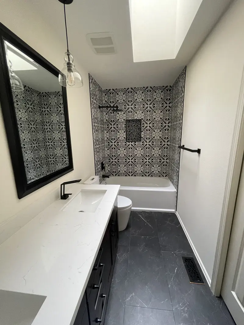 White counter with a black faucet under a black framed mirror, looking toward the patterned tile tub