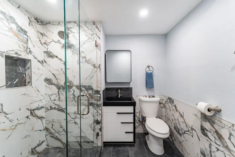 Remodeled bathroom with a frameless glass walk-in shower in white marble-look tile, a toilet and a white vanity with a black sink