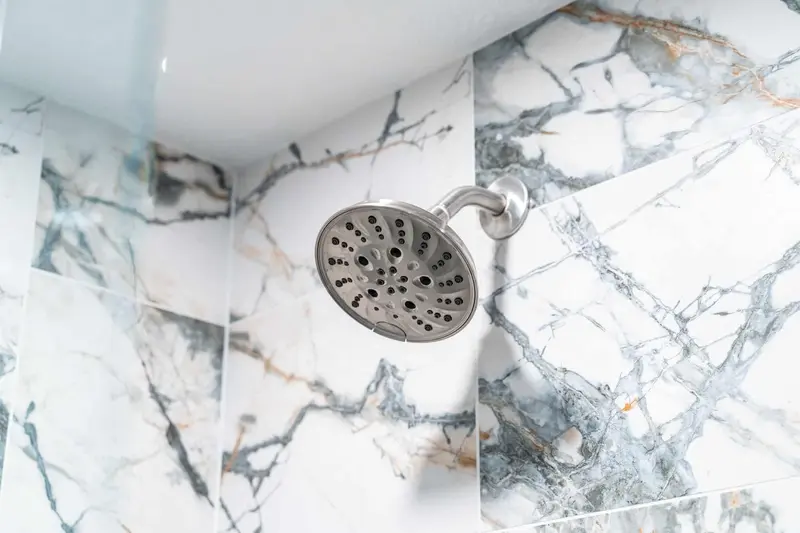 Brushed nickel shower head against white marble-look tile with gray and gold veins