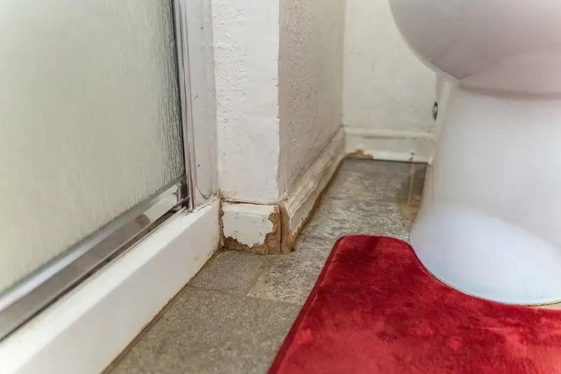 Worn trim and stained grout at the base of the old shower stall beside the toilet