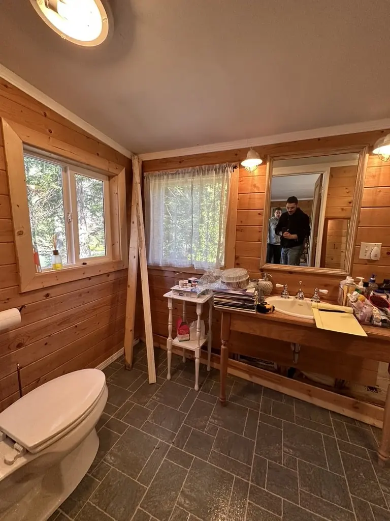 Pine paneled bathroom with a toilet, two windows and a table used as a vanity, before the remodel