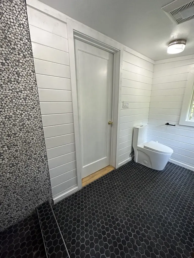 Bathroom door and toilet along white shiplap walls with a black hexagon tile floor