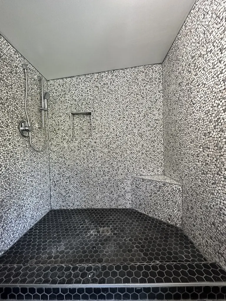 Walk-in shower tiled in black and white mosaic with a corner bench, a niche and a black hexagon floor