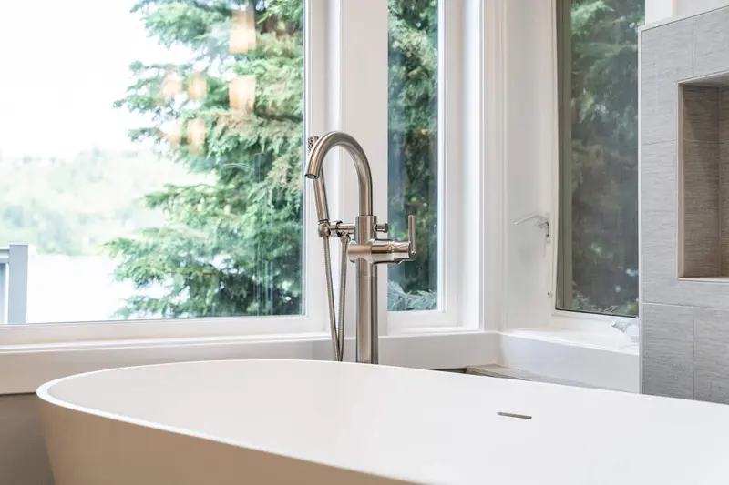 Floor-mounted tub filler beside a white freestanding tub, with trees outside the window