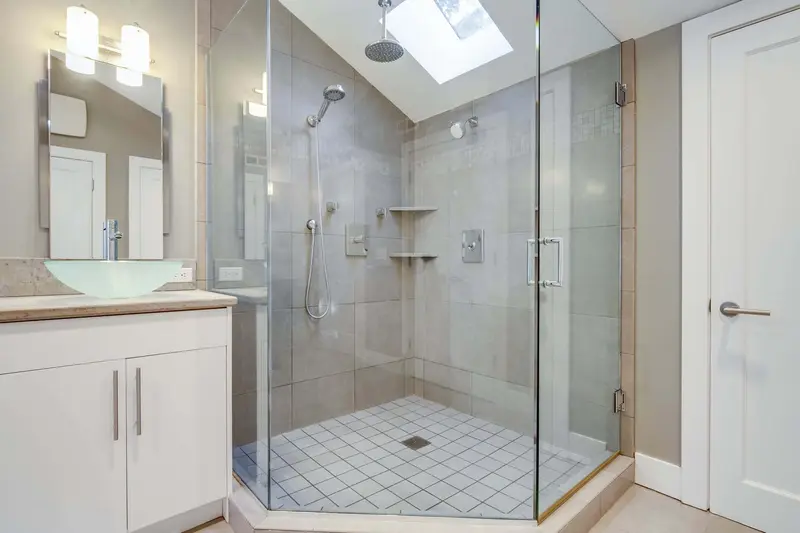 Old glass shower with a tiled floor beside a white vanity under a skylight