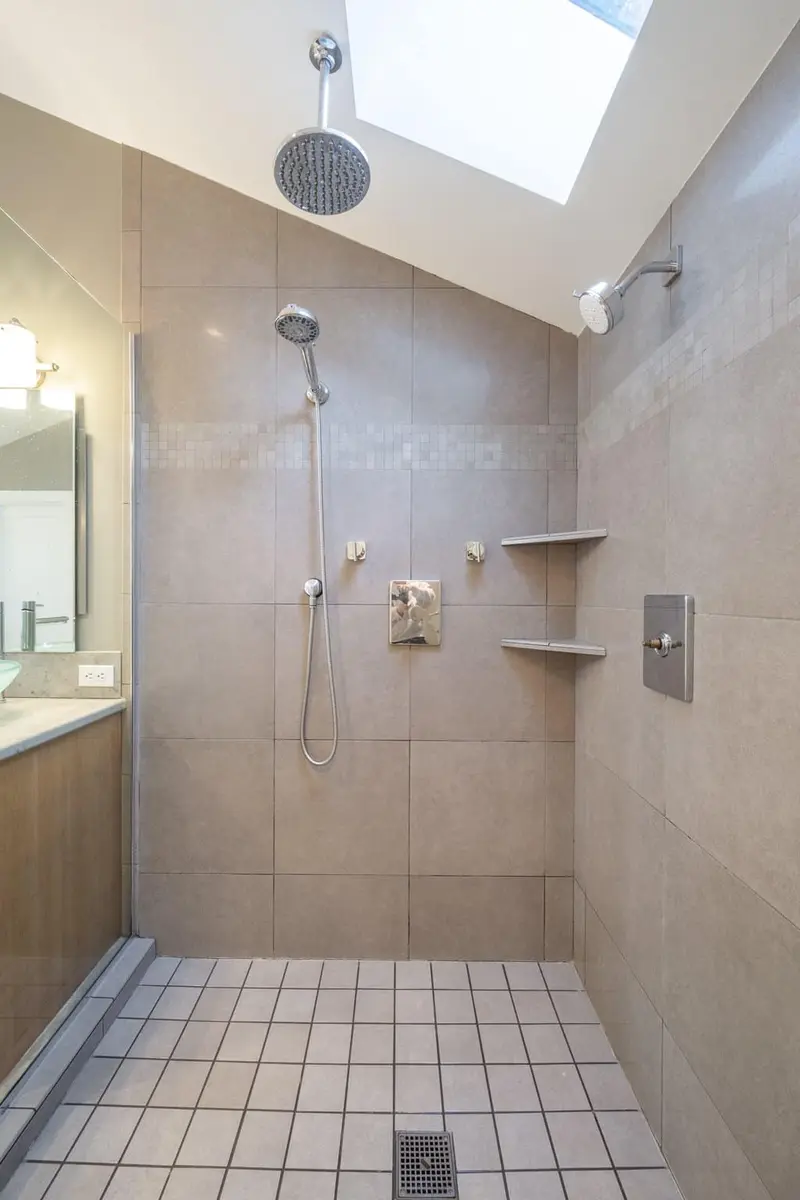 Inside the old shower with tan wall tile, corner shelves and a skylight above