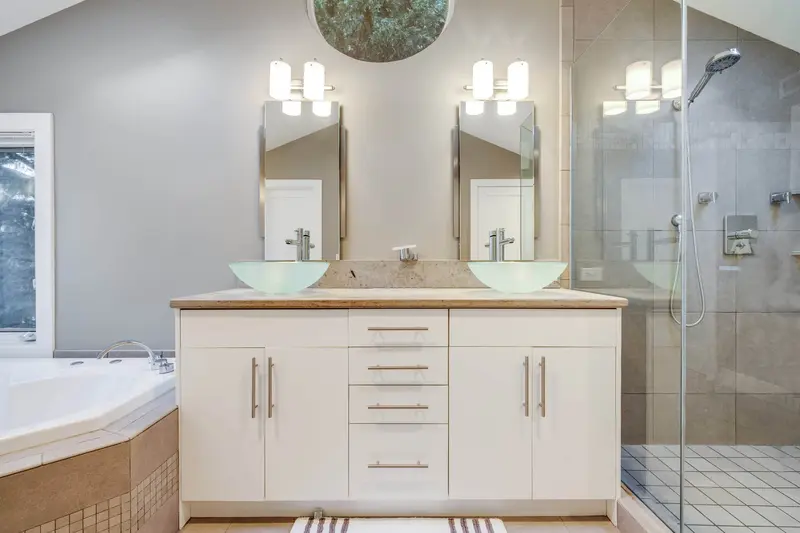 Old white double vanity with two glass vessel sinks and twin mirrors under a round window