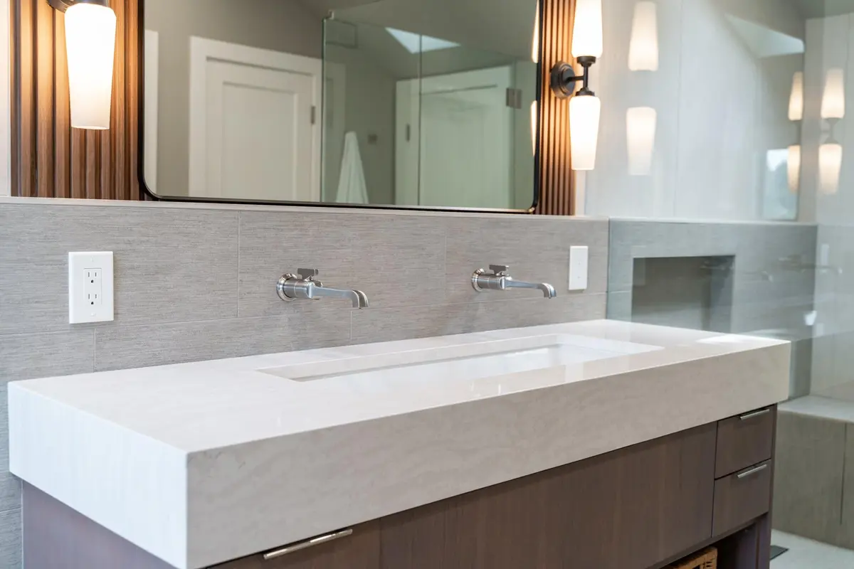 Wide white trough sink with two wall-mounted faucets on a floating wood vanity