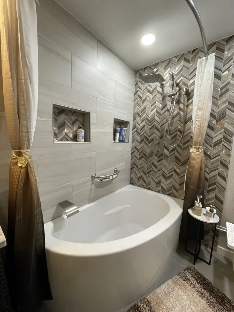 White corner soaking tub with chevron mosaic and light plank tile walls, two niches and a curved curtain rod