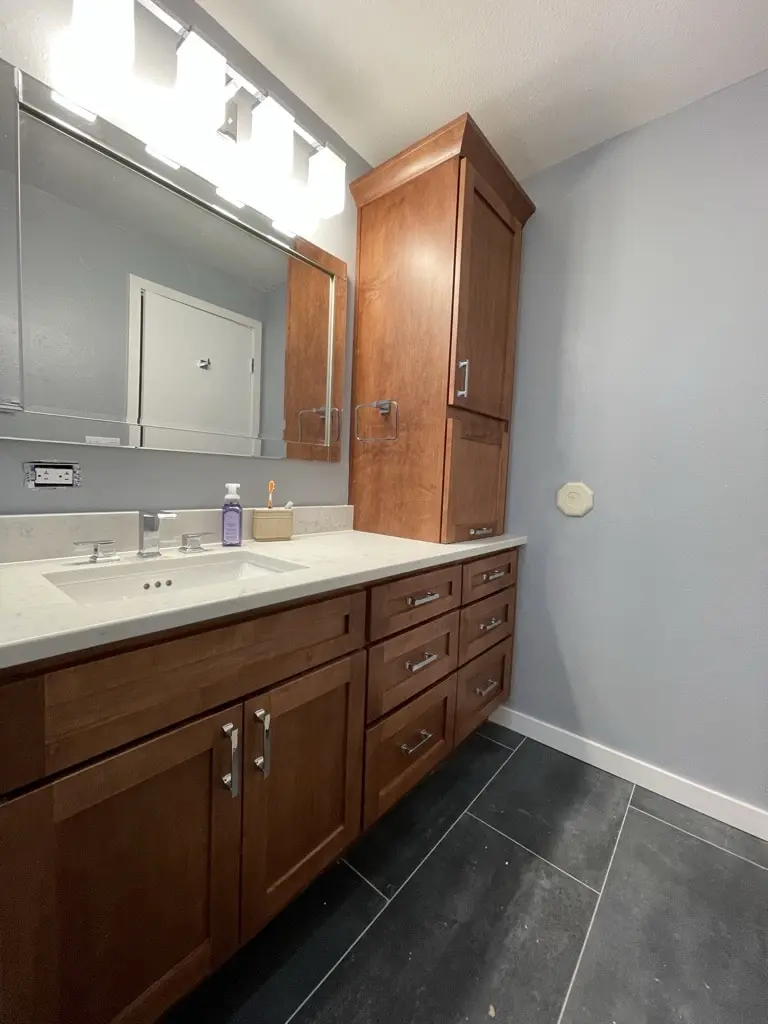 Light wood shaker vanity and tall linen tower on large dark floor tile beside a gray wall
