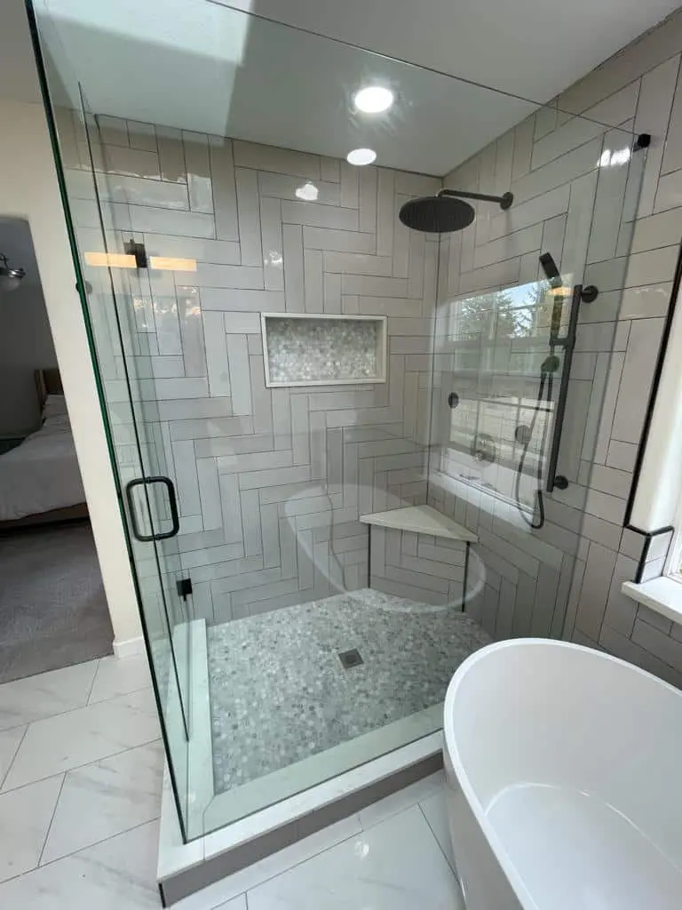 Frameless glass shower with gray herringbone tile, a rain head, a mosaic niche, a corner bench and a pebble floor