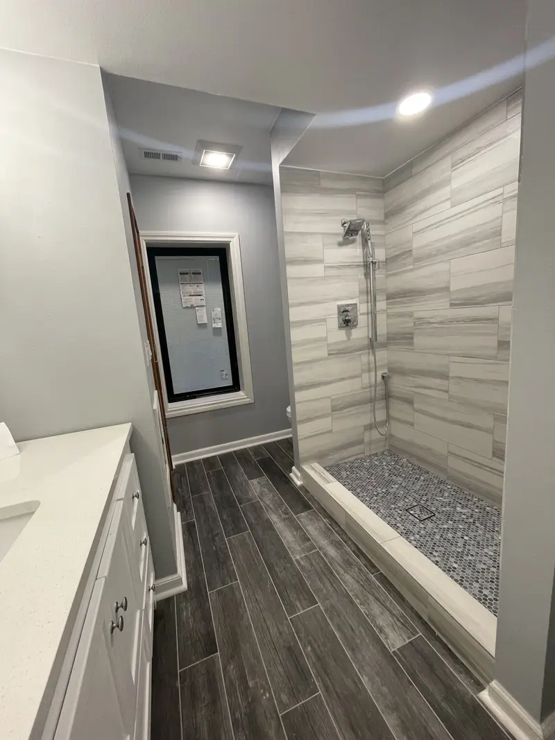 Open shower beside the white vanity, with plank tile running to a window at the end
