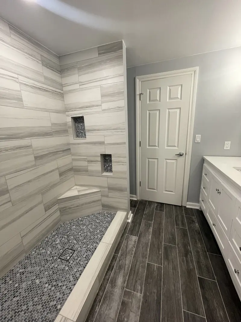 Walk-in shower tiled in long gray and white tile with a corner bench, two niches and a pebble floor, beside a white vanity