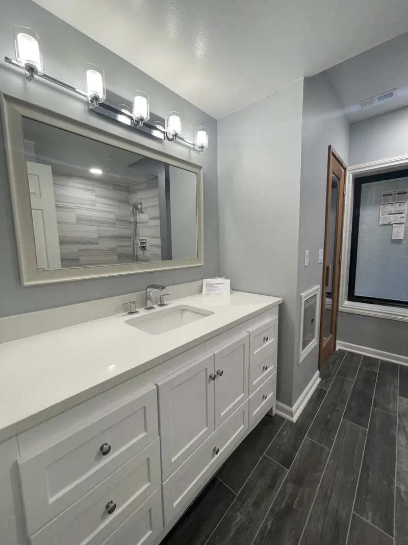 White shaker vanity with a quartz-look top, a framed mirror and a four-light bar over a dark wood-look tile floor
