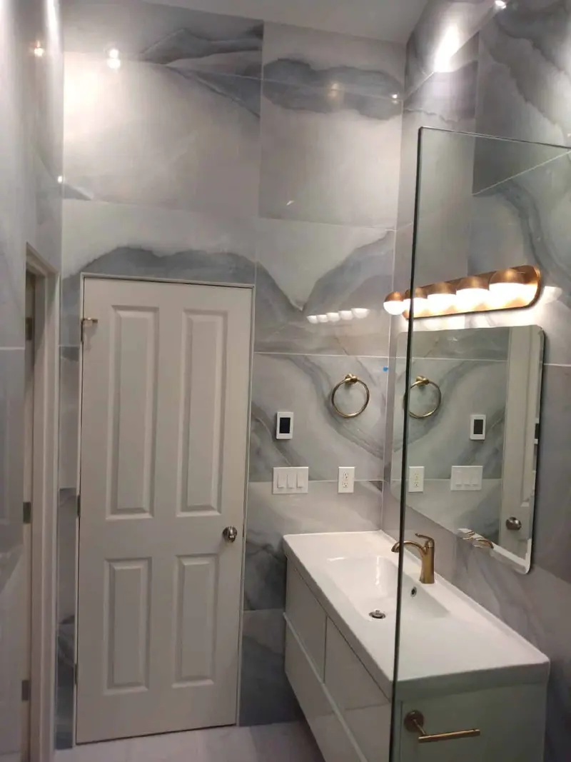 Floating white vanity with a brushed gold faucet and pulls, a gold light bar and towel rings on blue gray marble-look tile walls