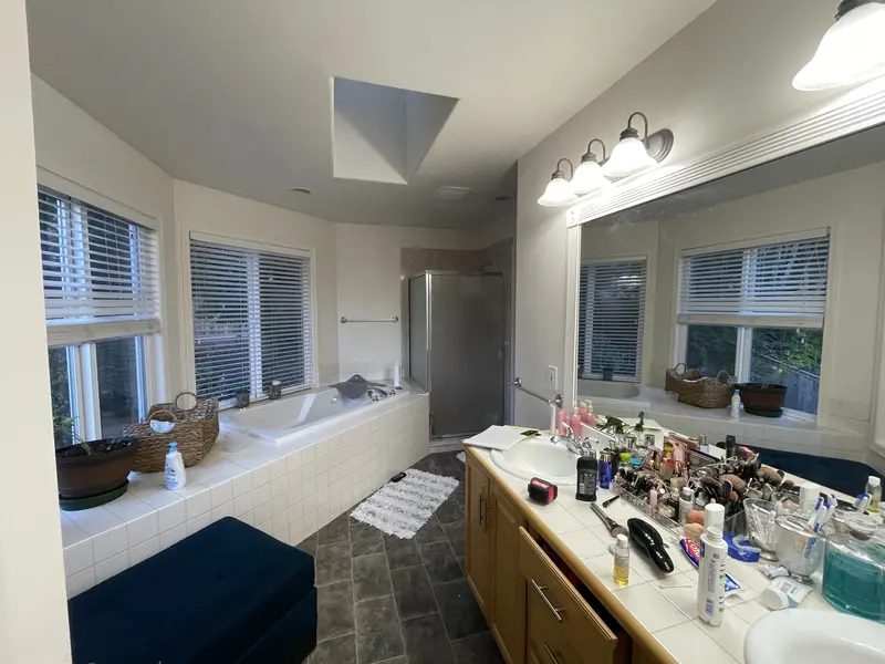 Bathroom before the remodel with a tiled tub deck in the window bay and an oak vanity covered in toiletries