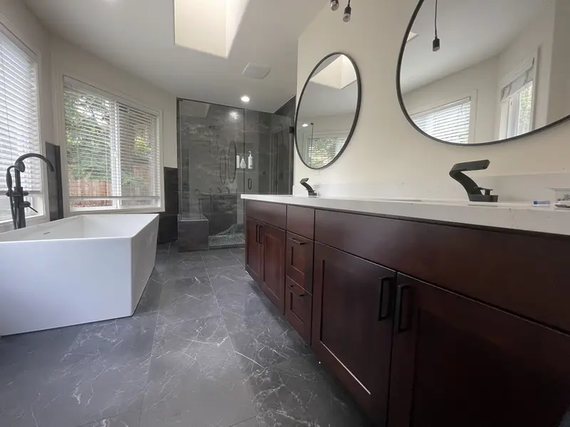 Remodeled bathroom with an espresso double vanity, round black mirrors, a freestanding tub and a glass shower
