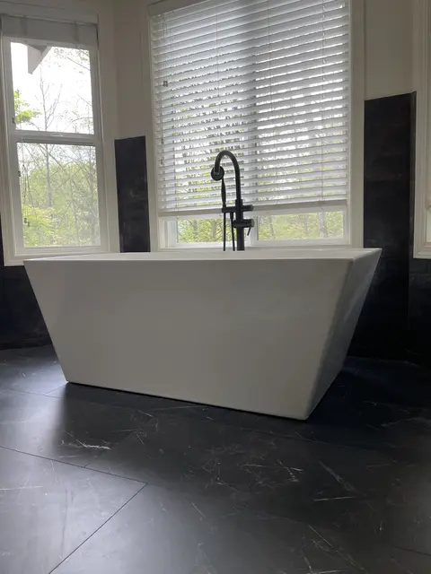 Freestanding white tub with a black floor-mounted filler in front of a window with dark wall tile