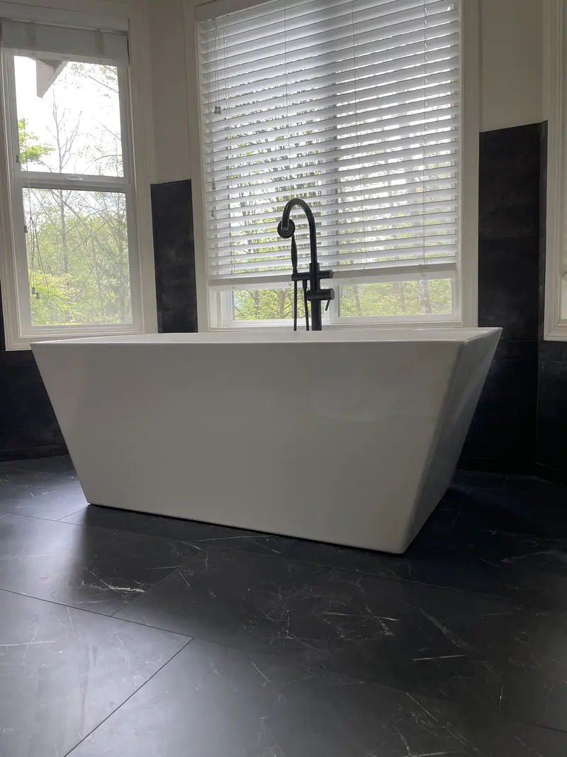 Freestanding white tub with a black floor-mounted filler in front of a window with dark wall tile