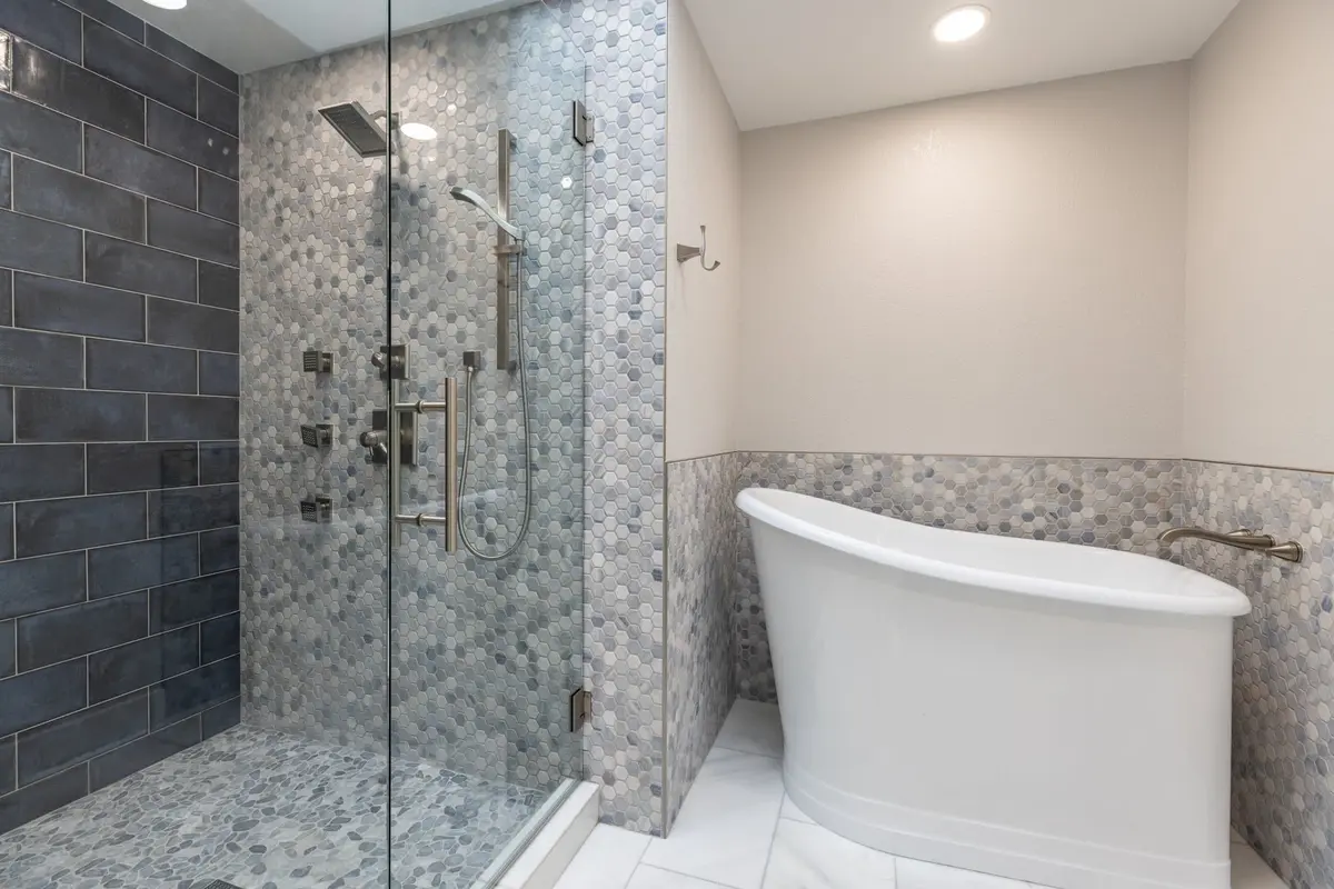 Walk-in glass shower with blue-gray hex mosaic beside a white freestanding soaking tub