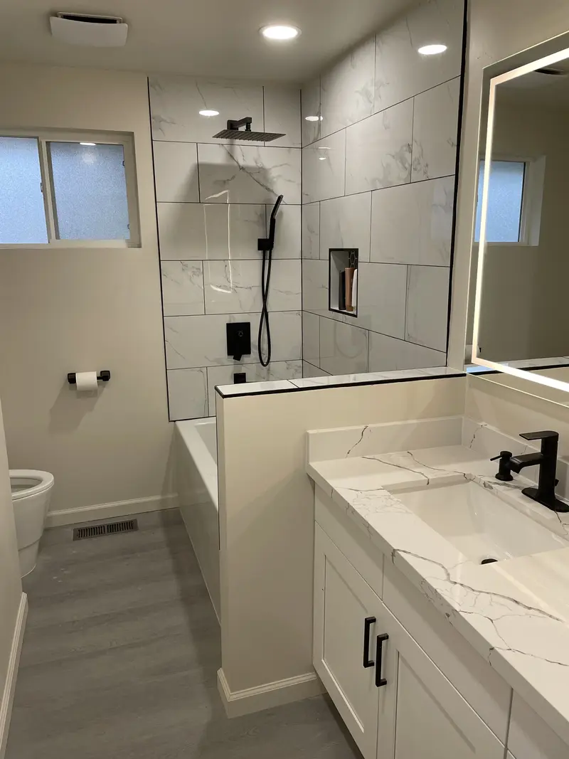 White shaker vanity with a veined top and black faucet next to a half wall and the tub