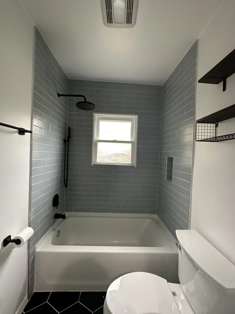 Tub with a gray tile surround, black shower rod and black rain head below a window