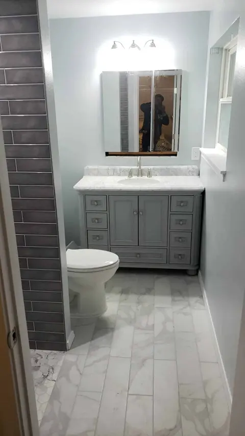 Remodeled bathroom with a gray vanity, white top, three-light fixture, charcoal tile wall and marble-look floor
