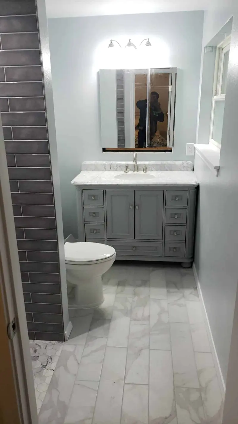 Remodeled bathroom with a gray vanity, white top, three-light fixture, charcoal tile wall and marble-look floor