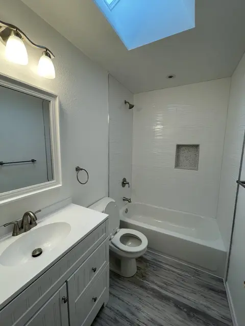 Remodeled bathroom with a gray shaker vanity, a framed mirror and a three-light fixture under a skylight, with the white tile tub beyond