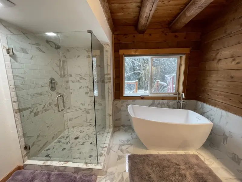 White freestanding tub under a window beside a frameless glass shower with marble-look tile