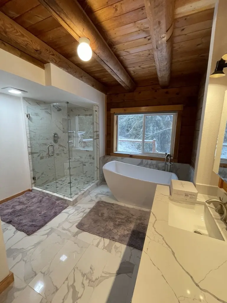 Remodeled log home bathroom with a frameless glass shower, a freestanding tub under the window and marble-look tile