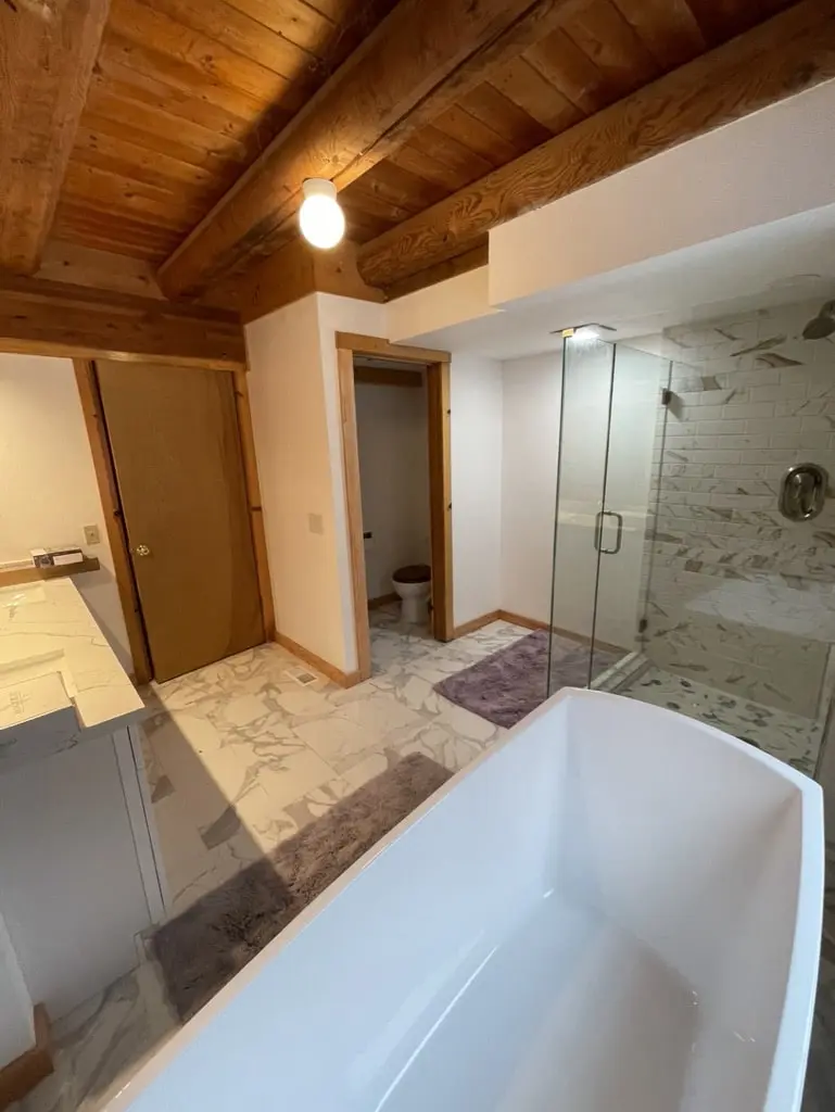 View past the freestanding tub to the glass shower and doors under the log beams