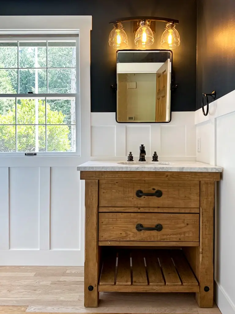 Powder room with a rustic wood vanity, black framed mirror and three-globe light against navy walls above white wainscoting
