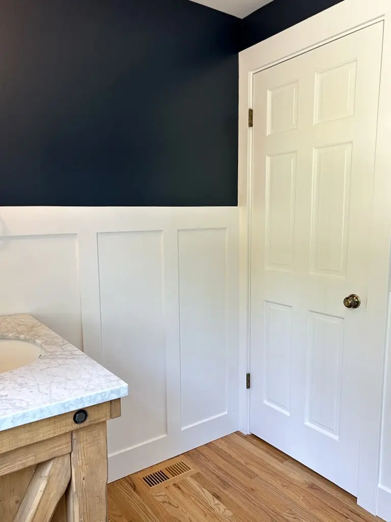 Corner of the powder room with white wainscoting, a white panel door and a light wood floor