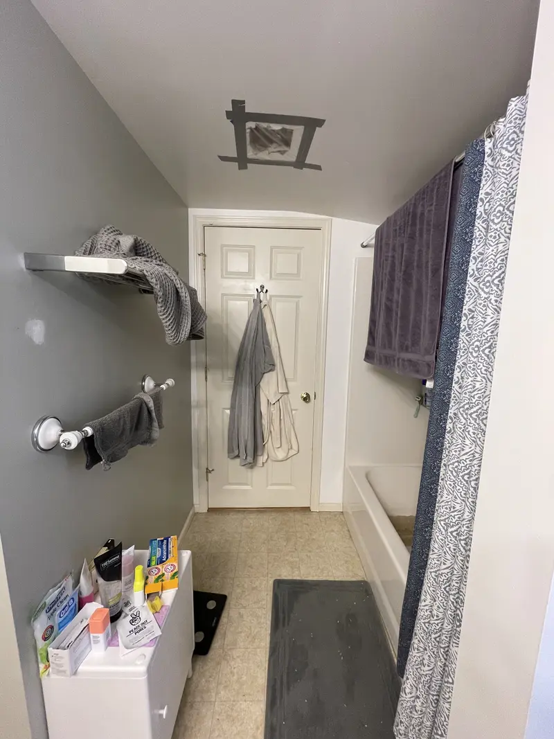 Bathroom before the remodel with a white tub behind a patterned shower curtain and a beige vinyl floor