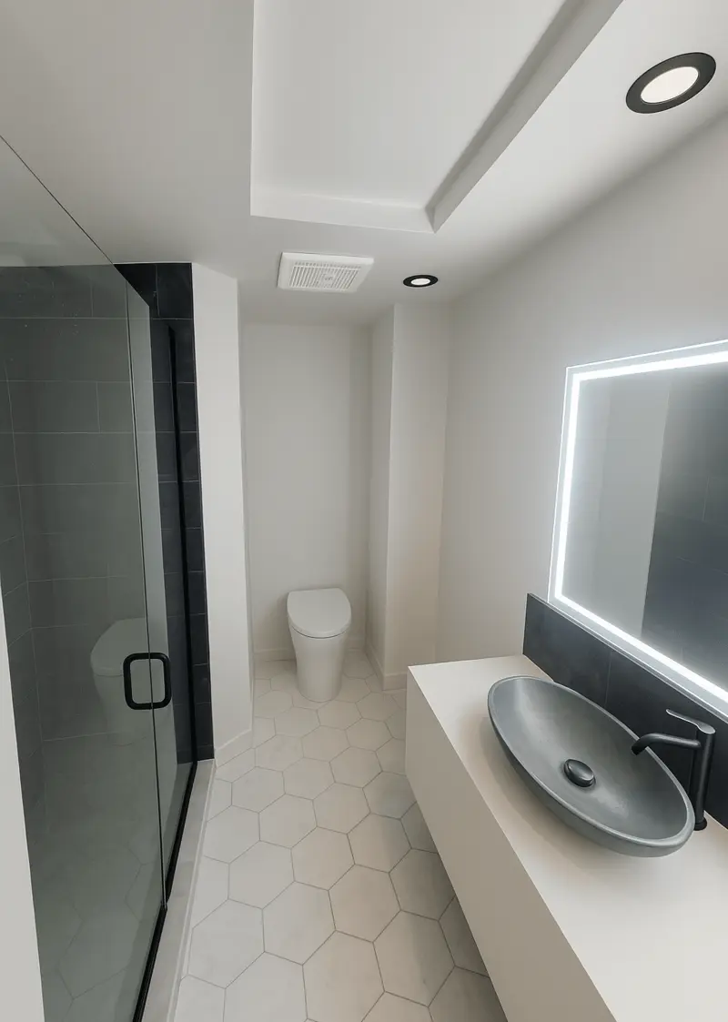 Finished bathroom with a glass walk-in shower in dark tile, a white vanity with a gray vessel sink, a lit mirror and white hexagon floor tile