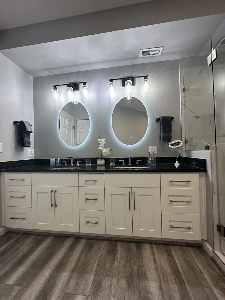White shaker double vanity with a dark counter and two lit oval mirrors under black sconces