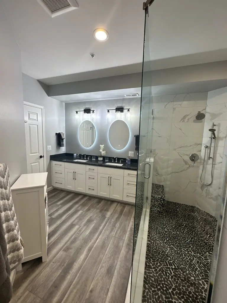 Bathroom with the walk-in shower in the foreground and a double vanity with lit oval mirrors beyond
