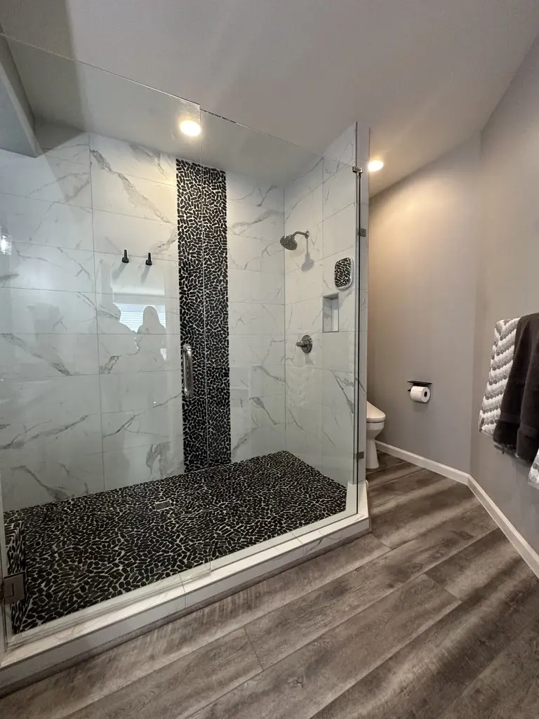 Large walk-in shower with marble-look tile, a vertical black pebble stripe and a pebble floor behind frameless glass