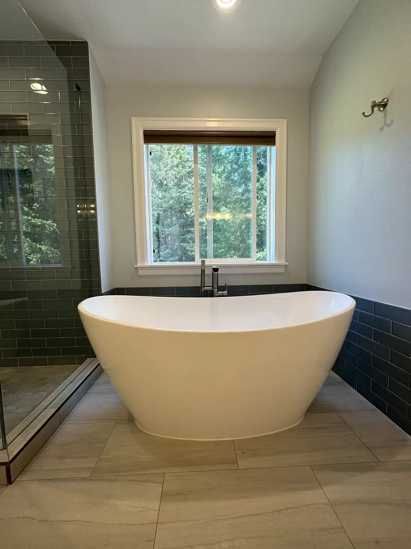 Freestanding white tub centered under a window, with dark tile wainscot behind it and a light wood-look floor
