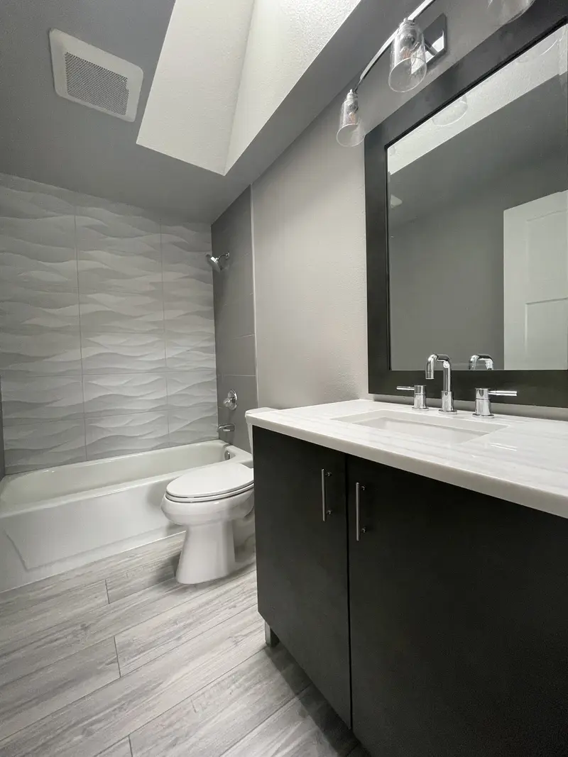 Bathroom with an espresso vanity and white top under a skylight, a toilet and a tub with a white wave tile wall