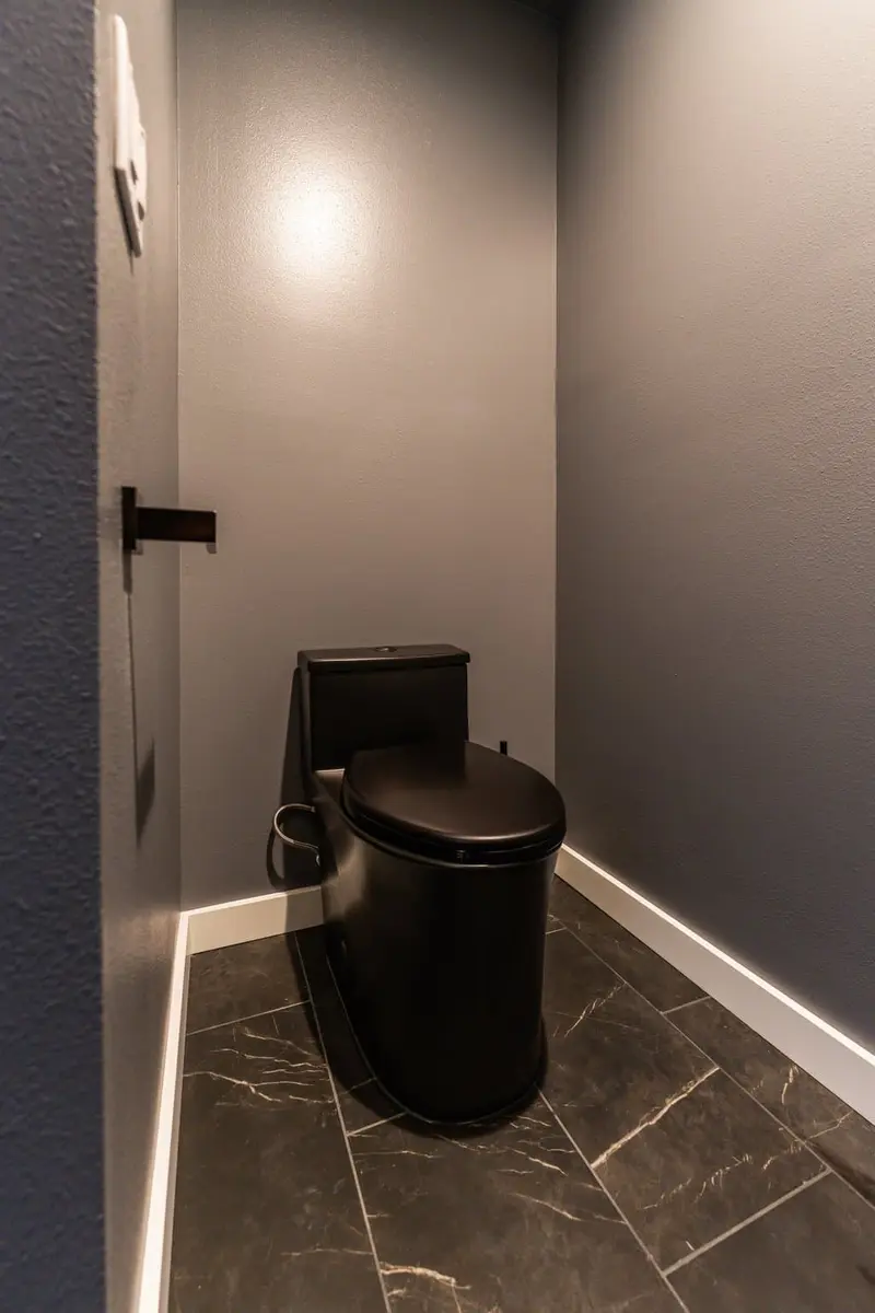 Black toilet in a gray-walled alcove on black marble-look floor tile with a lit baseboard