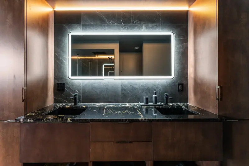 Wood double vanity with a black and gold stone-look top and a backlit mirror on black marble-look tile